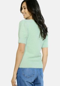 Šviesiai žalia pūkuota megztinis su trumpomis rankovėmis, raukšlėta apatine dalimi ir tekstūruotu audiniu. Atbulinės pusės vaizdas rodo prilipusį dizainą ir neformalų stilių.