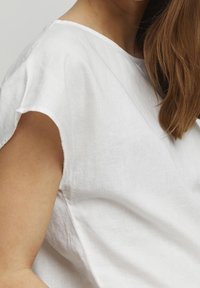 Weiße Leinenbluse mit lockerer Passform, kurzen Ärmeln und einer weichen Textur. Der Halsausschnitt ist rundlich und mit sichtbaren Nahtdetails versehen.