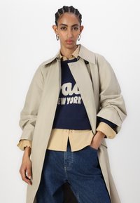 Trenci bej cu un guler structurat purtat peste un pulover bleumarin cu text alb și o cămașă crem cu nasturi. Blugi din denim închis la culoare.