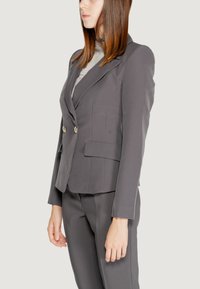 Blazer grigio su misura con doppi bottoni, design strutturato e tasche a patta. Il tessuto ha una texture morbida, che si abbina a pantaloni grigi aderenti.