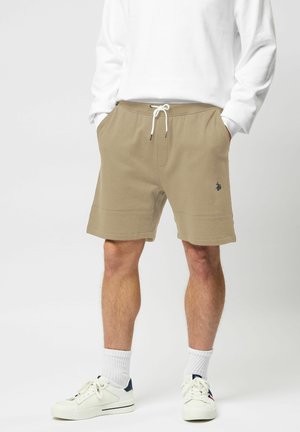 Mężczyzna w beżowych szortach z troczkami, białej koszuli z długim rękawem, białych skarpetkach i białych trampkach stojący z rękami w kieszeniach.