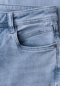 Jasnoniebieskie dżinsy z denimu, zbliżenie pokazujące przednią kieszeń, szlufkę na pasek i widoczne detale szwów na teksturowanym materiale.