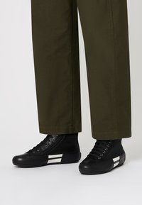 Sneakers alte nere con tessuto a trama e finitura in pelle, con strisce orizzontali bianche, abbinate a pantaloni verde oliva.