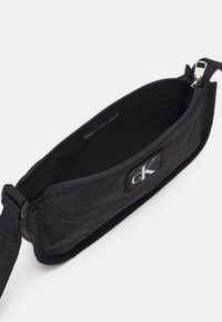 Černá látková crossbody taška se zipem, která má jemnou texturu a viditelné logo "CK". Vnitřek je podšitý černou látkou.