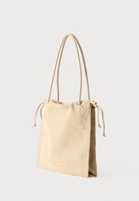 Τσάντα χειρός από σουέτ σε μπεζ απόχρωση με μακριές διπλές λαβές και πλαινά κορδόνια, σε όρθια θέση σε απλό λευκό φόντο.