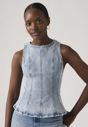 Žena s tmavou pokožkou a stiahnutými vlasmi, ktorá nosí kruhové náušnice a bezrukávový svetlomodrý džínsový top s detailmi šitia.