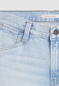 Jasnoniebieskie dżinsy z szerokimi nogawkami. Cechy obejmują kontrastowe przeszycia, zapięcie na guzik oraz metkę z napisem '94 Baggy Wide Leg.'