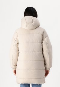 Manteau matelassé beige avec une capuche, présentant des sections matelassées et une coupe décontractée. Les poignets côtelés aux manches assurent une fermeture ajustée.