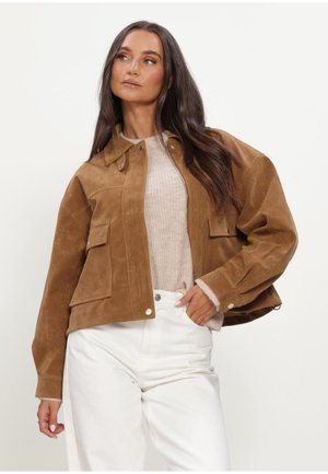 Vrouw draagt een bruine suède jas over een beige trui en witte broek, poseert met één hand in haar zak tegen een effen achtergrond.