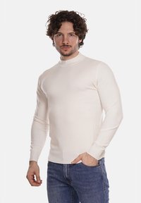 Maglione a collo alto a maniche lunghe color crema chiaro, realizzato in tessuto liscio, con un design aderente e polsini a coste, abbinato a jeans blu.