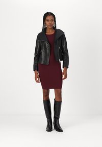 Crna kožna jakna sa širokim ovratnikom, uska bordo rebrasta haljina i crne čizme do koljena s sjajnom teksturom. Jednostavna pozadina.