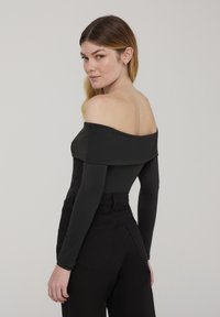 Top nero a spalle scoperte con maniche lunghe e texture liscia, abbinato a pantaloni neri a vita alta con tasche posteriori e design aderente.