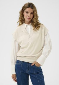 Beige ujjatlan pulóver, gallérral, gombos záródással. Fehér, hosszú ujjú inggel viselve, amelynek texturált mandzsettái vannak, és kék farmernadrággal.