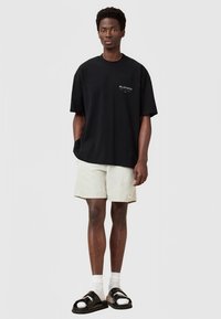 Čierne nadmerné tričko s malým logom, v kombinácii s svetlo béžovými šortkami, bielymi ponožkami a čiernymi sandálmi, stojace na pozadí bez vzoru.