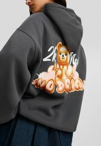 Sudadera gris con un gráfico de oso colorido y texto estilizado en la parte trasera, con una tela suave y texturizada y una gran capucha.