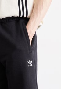 Čierne bavlnené teplákové nohavice s elastickým pásom a bočnými vreckami. Biele logo Adidas a podpisový detail troch pruhov pri páse.