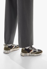 Sneakers i svart, grått och olivgrönt med texturerade paneler, en kraftig sula och djärv text "LE BLANCHE" på hälen. Matchas med grå byxor.