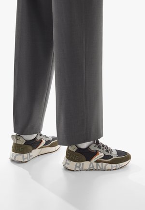Sneakers neri, grigi e oliva con pannelli testurizzati, una suola spessa e la scritta audace "LE BLANCHE" sul tallone. Abbinati a pantaloni grigi.