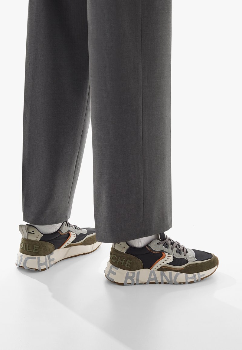 Sneakers i svart, grått och olivgrönt med texturerade paneler, en kraftig sula och djärv text "LE BLANCHE" på hälen. Matchas med grå byxor.