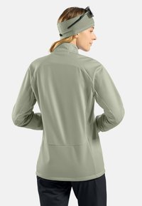 Eine Frau trägt eine hellgrüne Jacke und ein Stirnband mit Sonnenbrille auf dem Kopf, von hinten gesehen vor einem einfarbigen Hintergrund.