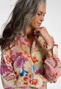 Kvinde med langt gråt hår iført en farverig blomsterprintet skjorte med knapper, kigger ned med hånden hvilende tæt ved ansigtet.