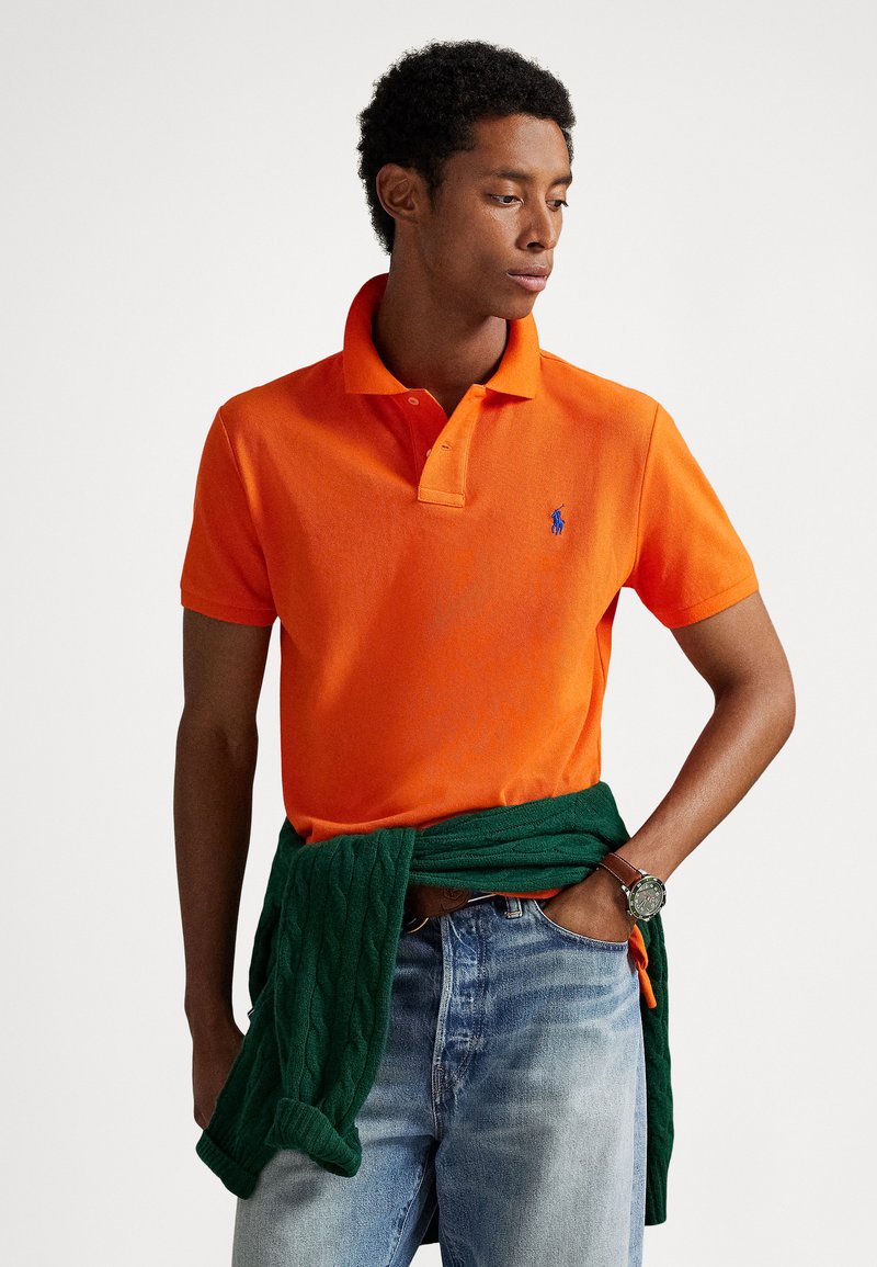 Polo orange avec col et logo bleu, associé à un jean bleu clair et un pull vert en maille torsadée noué autour de la taille.