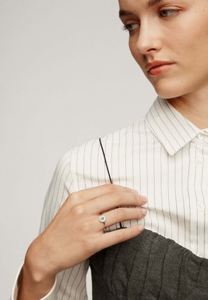 Anillo de plata con una piedra azul redonda, usado en una mano. La persona lleva una camisa blanca a rayas sobre una prenda gris texturizada.