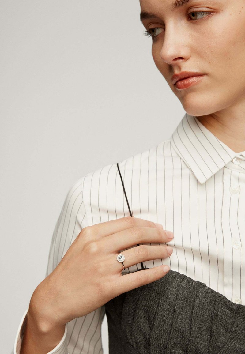Anillo de plata con una piedra azul redonda, usado en una mano. La persona lleva una camisa blanca a rayas sobre una prenda gris texturizada.