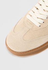 Beżowe zamszowe sneakersy o teksturowanym wykończeniu, kremowe sznurówki i podeszwa z gumy o wzorze jodełki.
