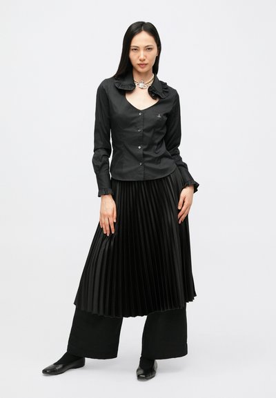 Melnā blūze ar garām piedurknēm, volānu apkakli un pogām, saskaņota ar melnu kroku svārku un melnām kurpēm, attēlota uz balta fona.