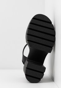 Sandale noire avec semelle en caoutchouc épaisse et rainurée et talon bloc, positionnée avec la semelle face à un fond d'angle blanc.