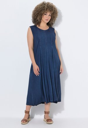 Kobieta z kręconymi włosami, ubrana w niebieską, bezrękawnikową sukienkę midi i brązowe sandały na koturnie z paskiem wokół kostki, stoi na białym tle.
