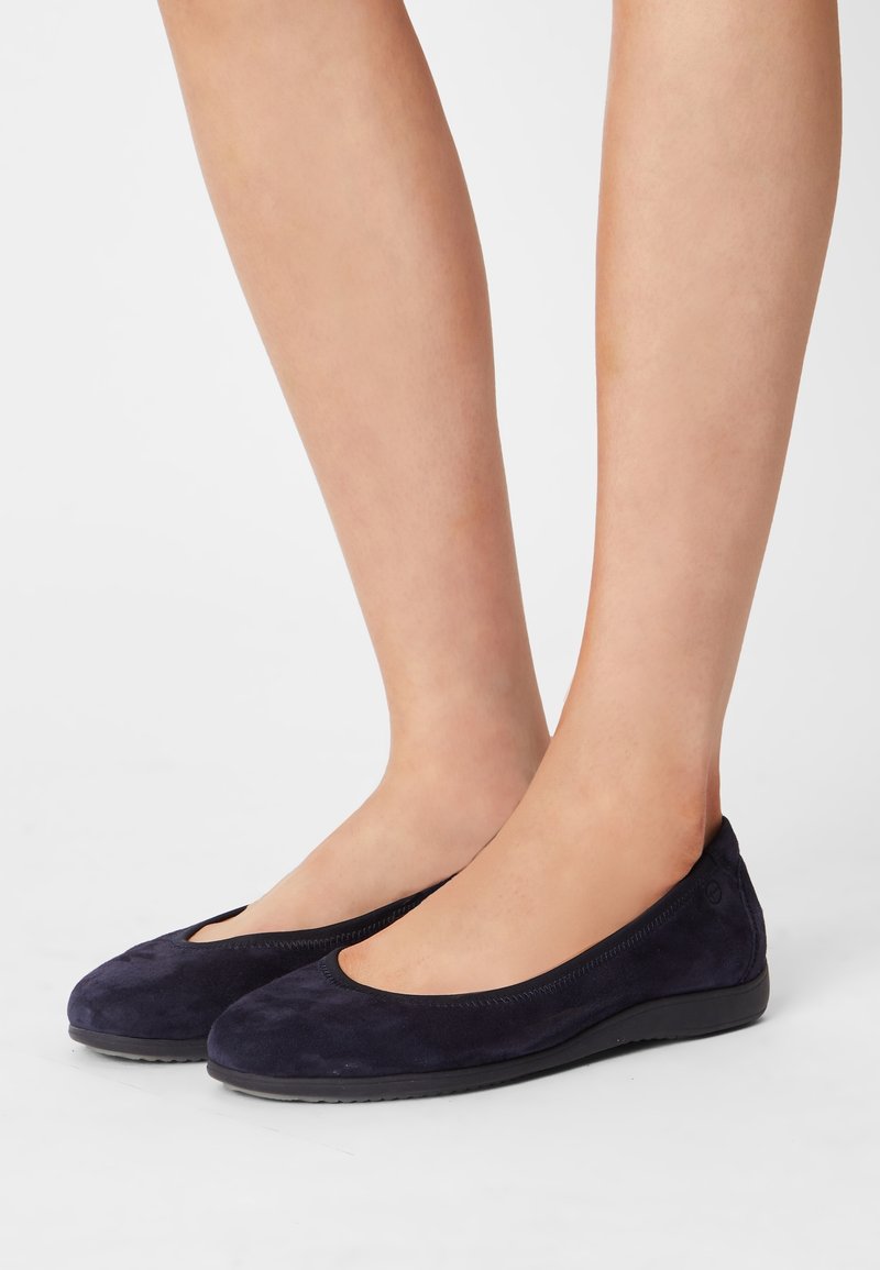 Paire de jambes nues portant des ballerines simples en daim bleu foncé avec des bouts arrondis et des semelles basses noires sur un fond clair uni.