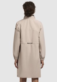 Impermeabile beige con collo alto, polsini elastici e vita cinte. Texture liscia e design minimalista, dotato di tasca sul retro.