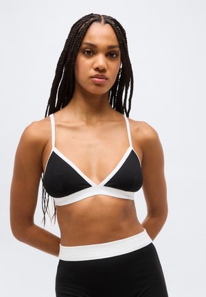 Žena s dlhými vrkočmi, oblečená v čiernobielom trojuholníkovom bralette a čiernych nohavičkách s vysokým pásom na jednoduchom pozadí.