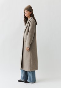 Lange lichtbruine wollen jas met een notched kraag, twee voorzakken en zwarte knopen, gecombineerd met wijde blauwe jeans en een bruine gebreide beanie.