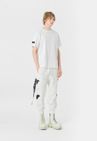 Camiseta blanca de algodón con un parche cuadrado; pantalones de chándal blancos a juego con acentos gráficos negros y cordones; botas de combate de color claro.