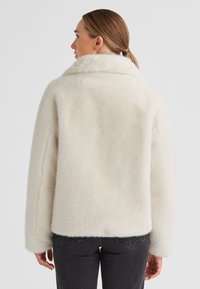 Flauschige, cremefarbene, oversized Jacke mit breitem Kragen, herabgesetzten Schultern und geradem Saum, kombiniert mit dunkelgrauen Hosen.