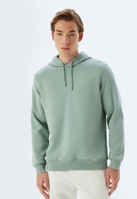 Sudadera con capucha de color verde claro, de corte relajado, con puños y dobladillo acanalados. Textura suave, diseño minimalista y cordón en la capucha.