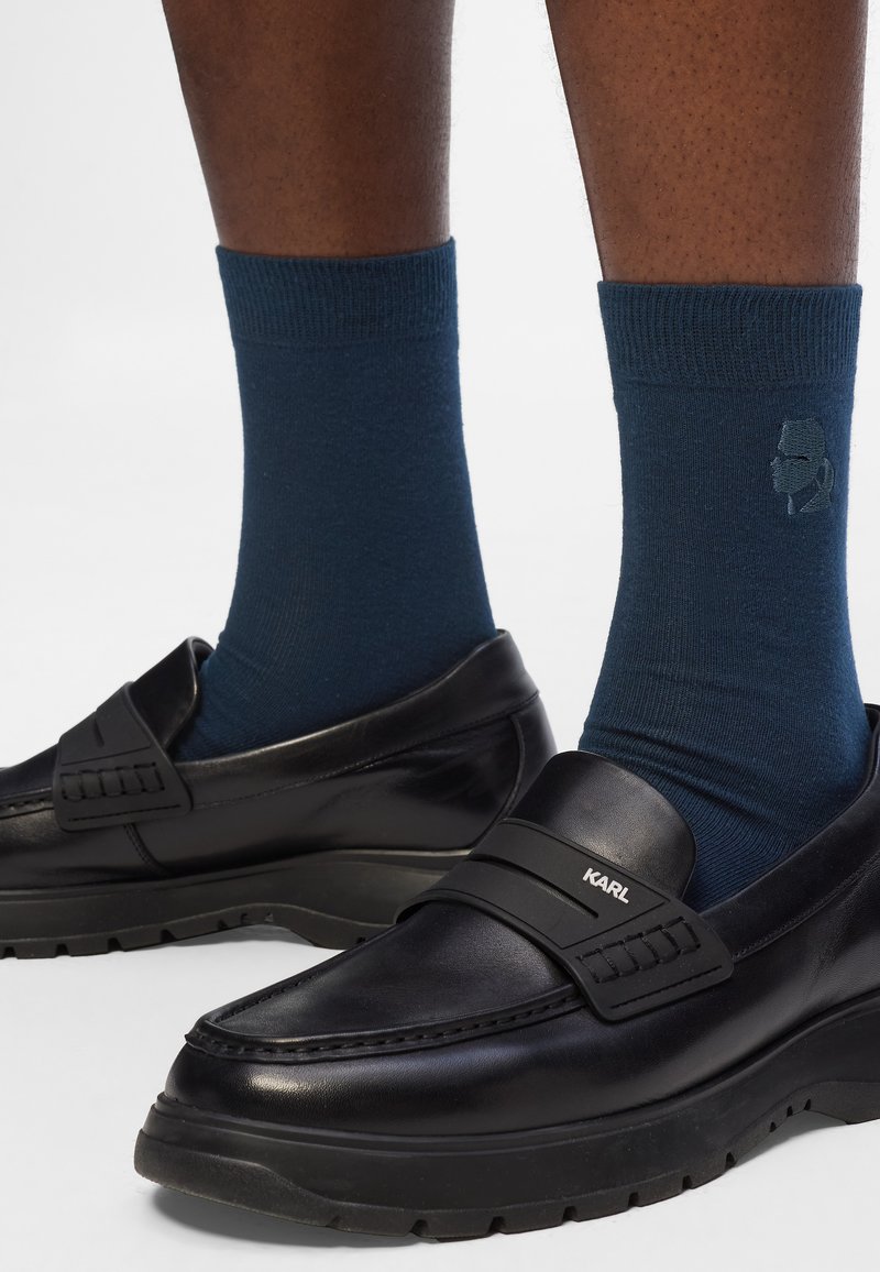 Mocassini in pelle nera con una fascia prominente etichettata "KARL," abbinati a calzini blu navy con un motivo logo, su uno sfondo neutro.