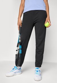 Person som har på seg svarte joggebukser, hvite og blå sportssko, holder en tennisracket i den ene hånden og en tennisball i den andre.
