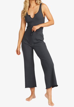 Femme portant un débardeur côtelé gris foncé avec une encolure en encoche et un pantalon large à cordon assorti, debout pieds nus.