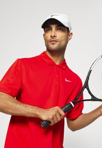 Rød polo shirt med krave og et Nike-logo. Figuren holder et sort tennisracket med struktureret greb. Hvid cap med perforeringer.