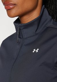 Mornarska zip-up jakna iz gladkega materiala, z visokim ovratnikom in belim logotipom na prsih. Podrobnosti zadrge so prikazane v bližnjem posnetku.