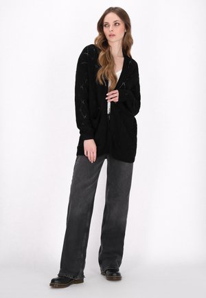 Cardigan nero in maglia aperta con scollatura a V, maniche ampie e vestibilità morbida, abbinato a jeans in denim grigio a vita alta e stivali neri.