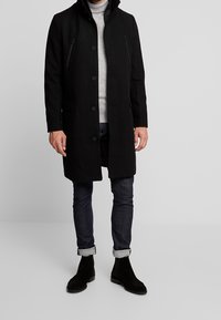 Schwarzer Wollmantel mit hohem Kragen, vorderen Knöpfen und zwei Reißverschlusstaschen auf der Brust. Kombiniert mit einem grauen Rollkragenpullover, dunklen Jeans und schwarzen Wildlederstiefeln.