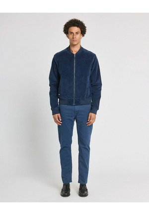 Homme debout face à nous, portant une veste zippée bleu marine, un pantalon bleu et des chaussures noires sur un fond clair uni.