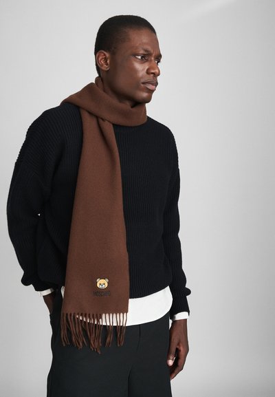 Sciarpa di lana marrone con estremità frangiate, caratterizzata da un logo di orso ricamato e il testo "Moschino". Indossata sopra un maglione nero con texture.
