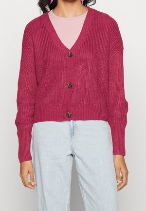 Kvinde iført en mørkerød strikket cardigan med tre knapper over en lyserød skjorte og lyseblå jeans.