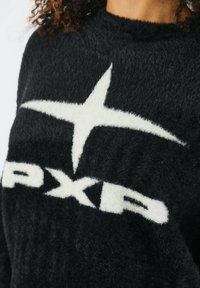 Schwarzer Pullover aus einem flauschigen Material mit einem großen weißen Stern und dem "PXR"-Logo. Runder Ausschnitt, weiche Textur und lockere Passform.